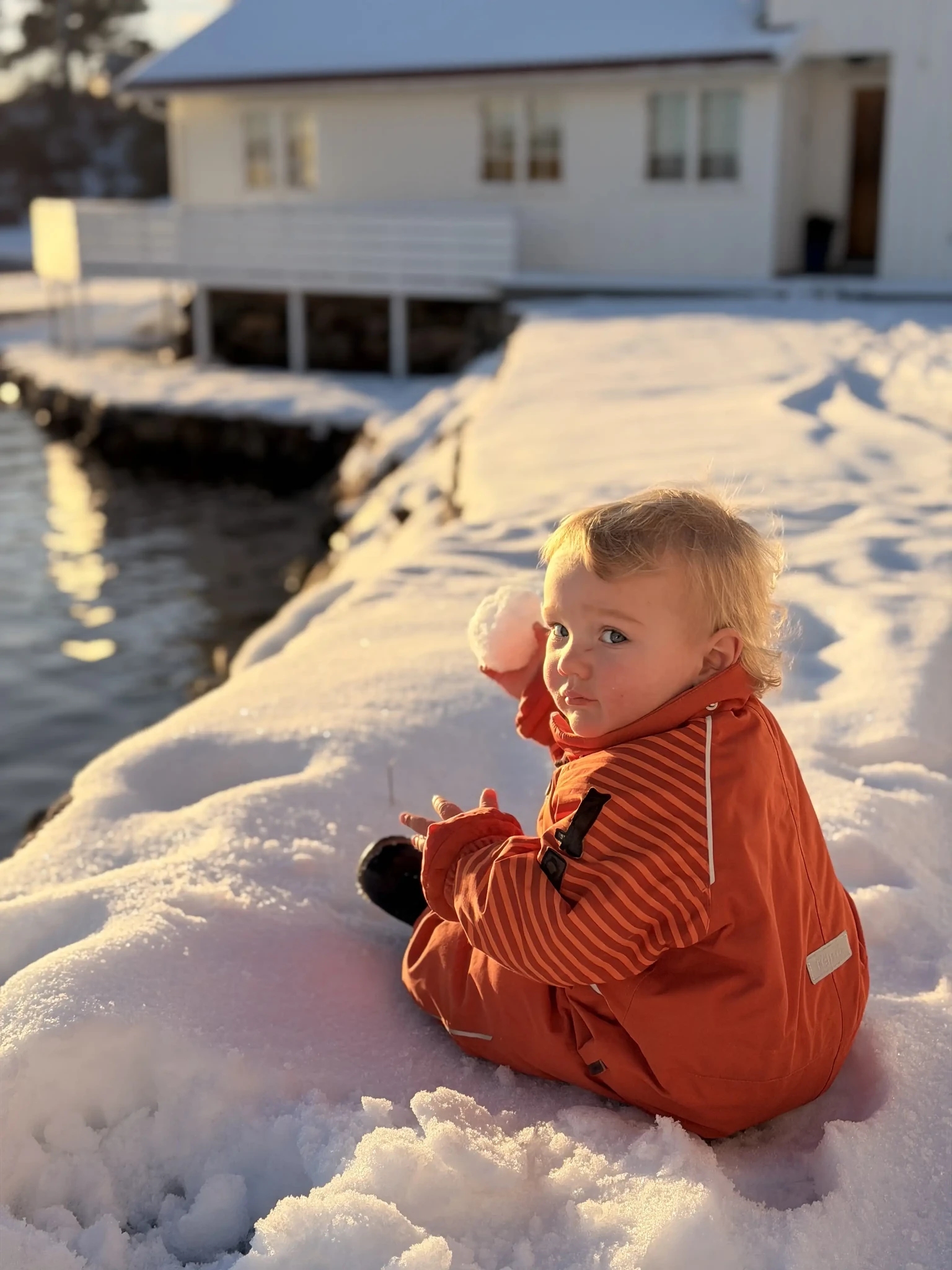 A child in an orange snowsuit holding a snowball beside the water and a white house.