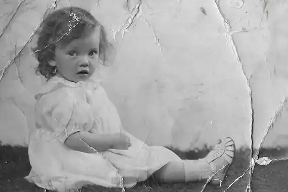 An old black-and-white photograph of a small child in a light dress, with creases and cracks across the paper.